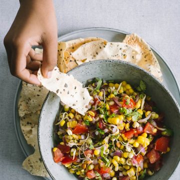 Hand holding cracker over a blue bowl of sprouts and corn salsa