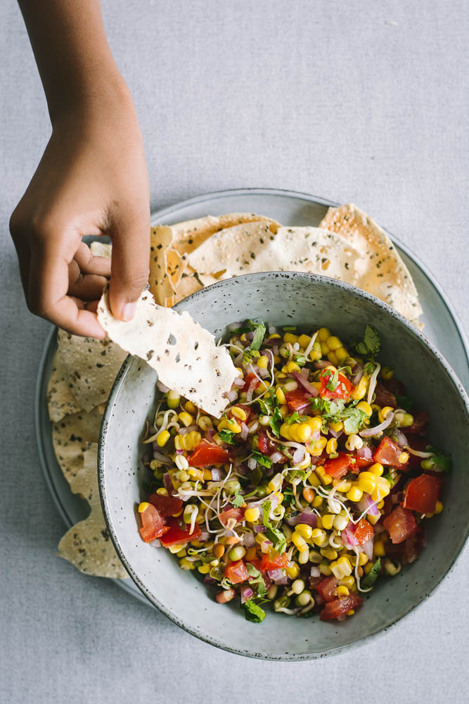 Hand holding cracker over a blue bowl of sprouts and corn salsa