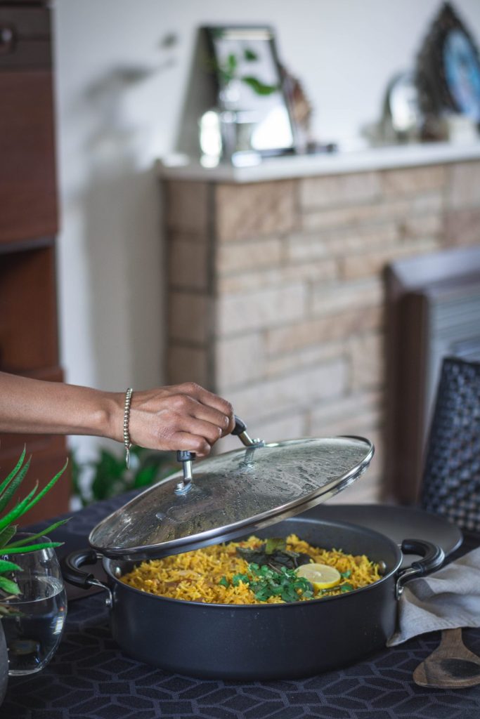 Hand lifting lid of large pan with chickpea biryani on dining table