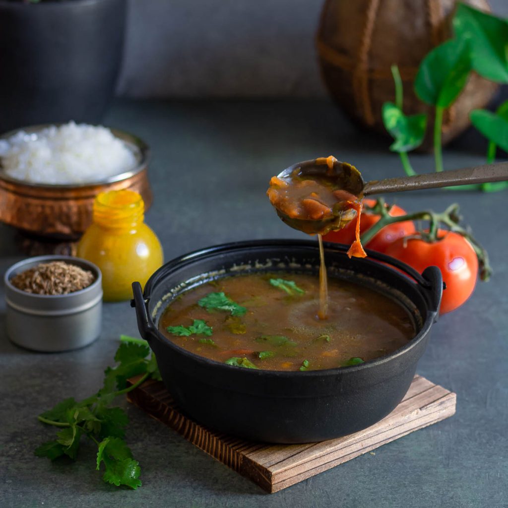 pouring indian tomato soup in a black bowl with ladle, spices, rice and fresh tomatoes in background