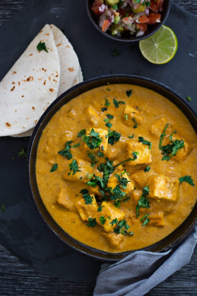 Indian cottage cheese curry served in black bowl with flatbreads and salad on the side