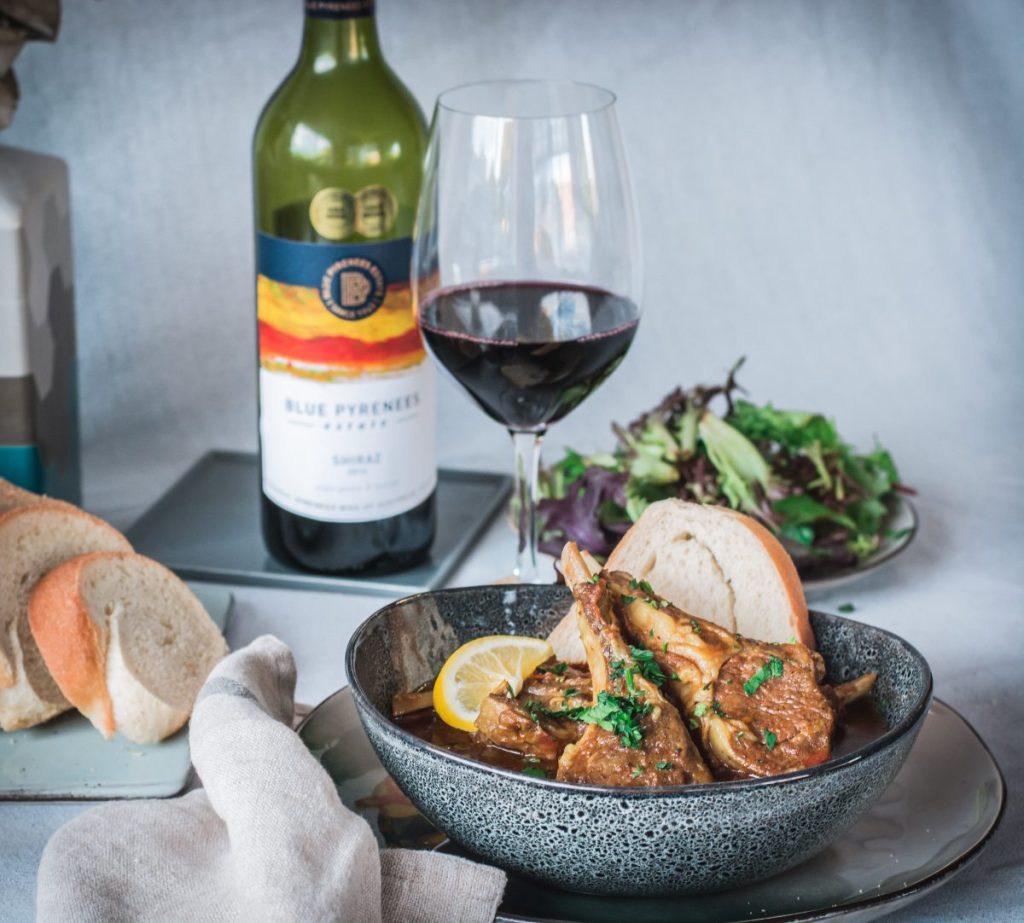 lamb chops curry in blue bowl with a glass of red wine and salad on the side