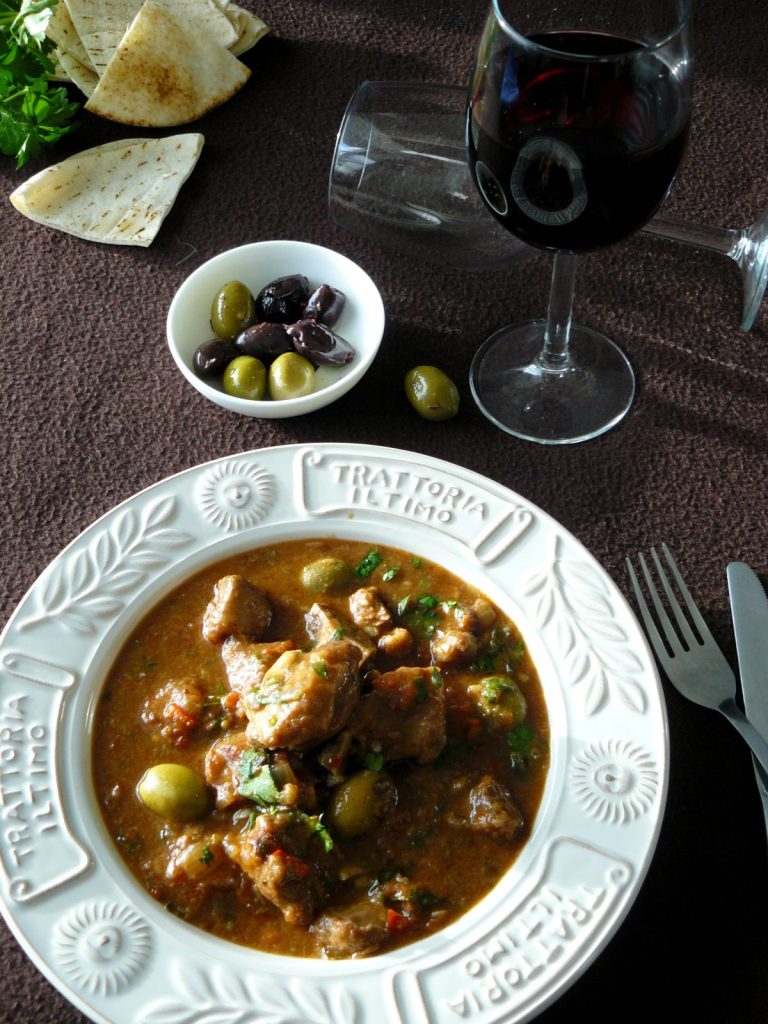 Italian lamb stew in white plate, a bowl of olives and a glass of red wine on the side