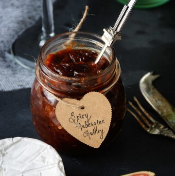 Aubergine chutney in small glass jar with a wedge of cheese and figs on the side