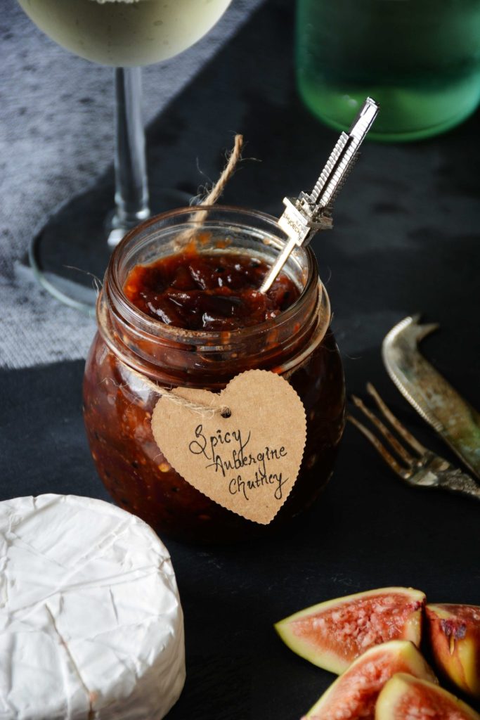 Aubergine chutney in small glass jar with a wedge of cheese and figs on the side