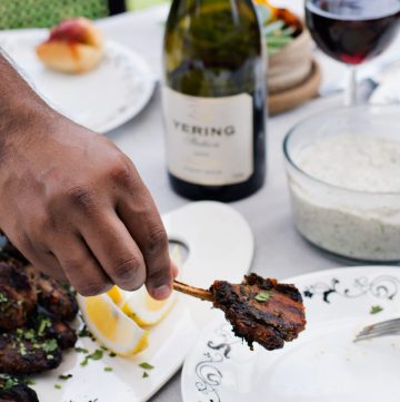 Hand holding a grilled lamb cutlet with a bottle and glass of red wine in the background