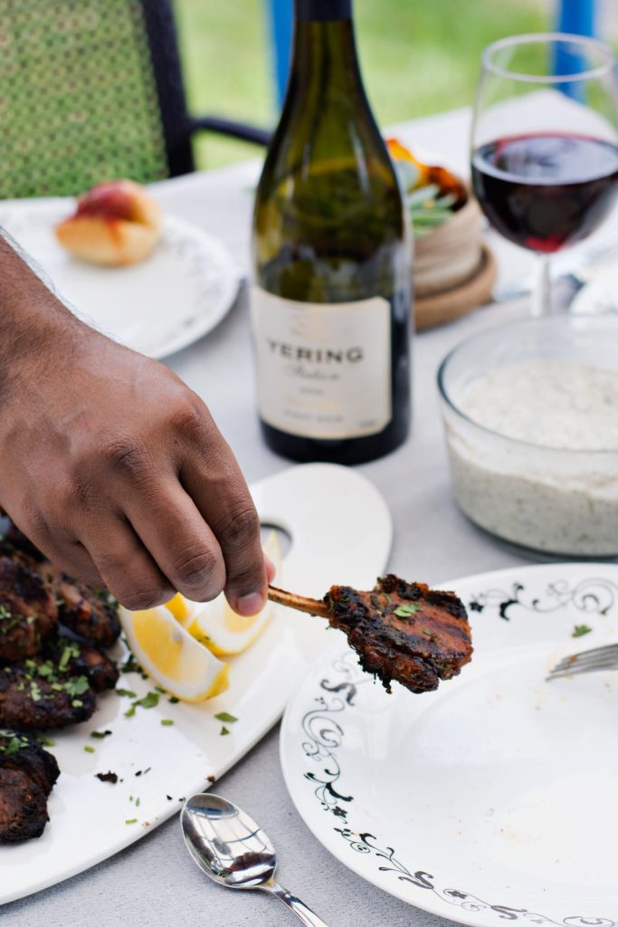 Hand holding a grilled lamb cutlet with a bottle and glass of red wine in the background