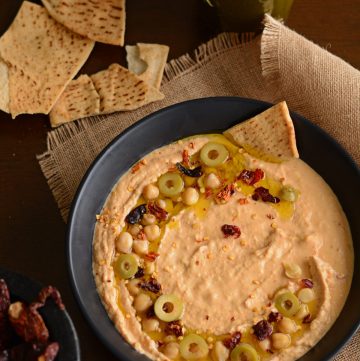 Chilli infused hummus garnished with sliced olives, chickpeas served in black bowl with pita bread on the side