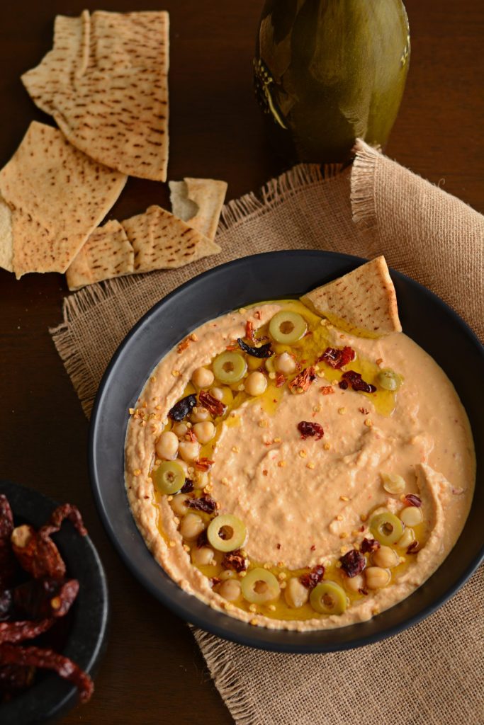 Chilli infused hummus garnished with sliced olives, chickpeas served in black bowl with pita bread on the side