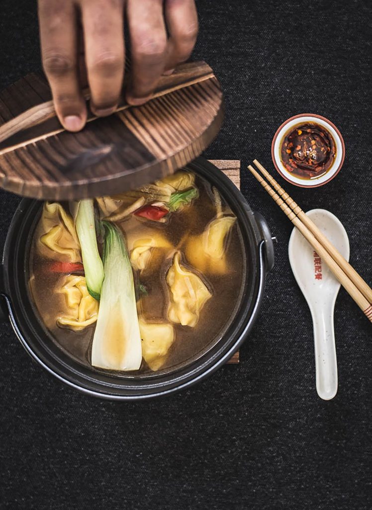 hand lifting lid of black bowl with chicken wonton soup