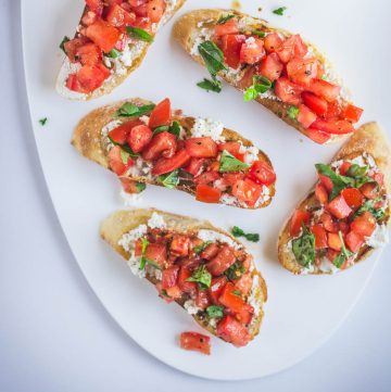 five slices of bruschetta with tomatoes and marinated feta on white platter